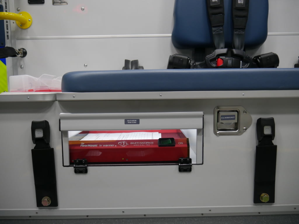 A SmithWorks Medical Inc. FloorMount IV warmer is contained in a clear-faced compartment under the squad bench.